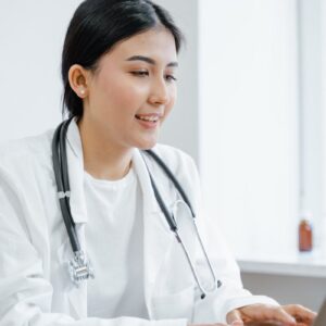 Asian female doctor in a white coat typing on a laptop during a telemedicine session.